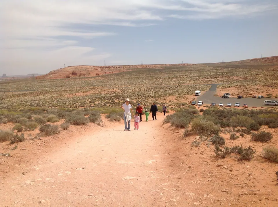 A woman in a white sun hat and three others, including a small girl in pink and a child in green, walk up the orange trail from the parking lot with the desert flats behind.