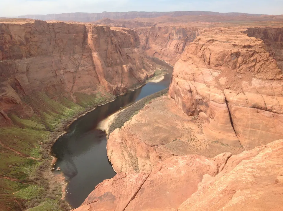 The Colorado curves around a huge sandstone point far below the rim, with green banks on the left and the far canyon walls beyond.
