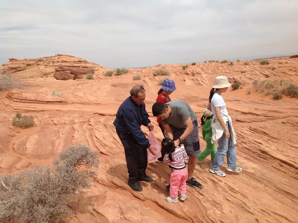 On a ledge of banded orange sandstone, an older man in a dark jacket helps a small girl while a man in grey bends beside them and a woman in a white hat and a child in green look on.