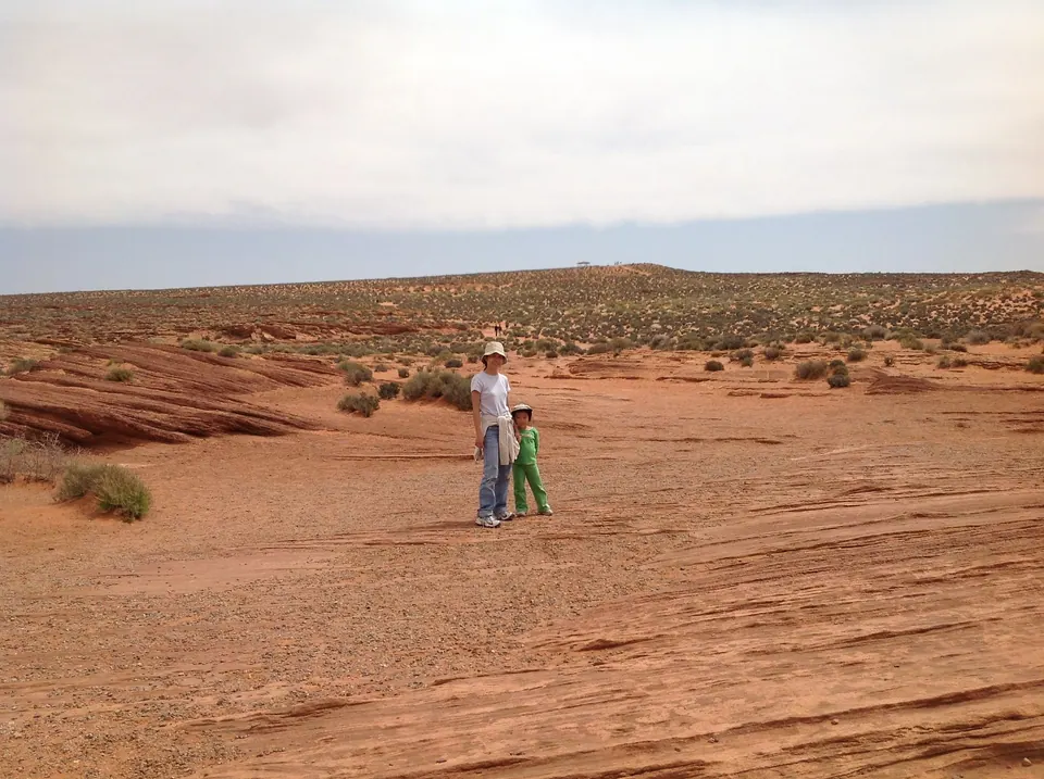 A woman in a white hat and a child in green stand on a wide flat of swirled red sandstone, with a low horizon and cloudy sky.