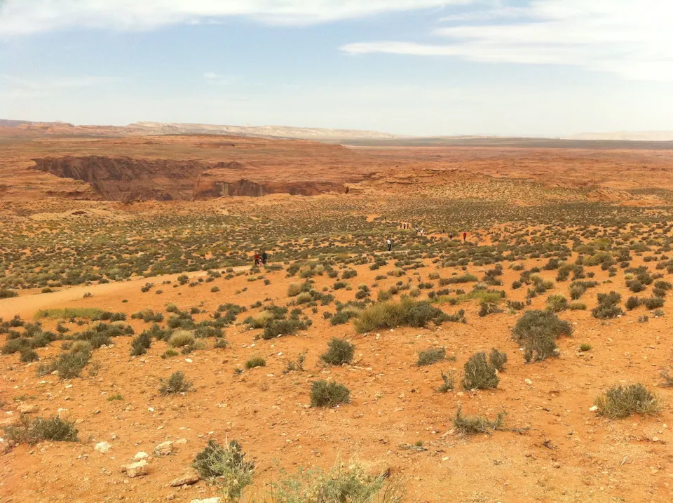 A view back over orange desert and scrub toward a gash in the sandstone, with tiny walkers on the trail.