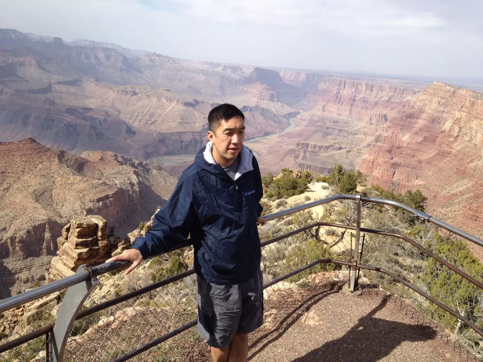 A man in a navy rain jacket stands at a railing with the layered canyon and the Colorado River winding far behind him.