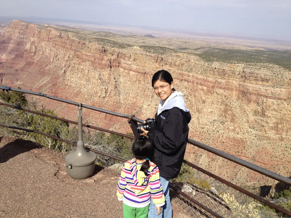 A smiling woman in a black jacket with a white hoodie collar holds a camera at the railing beside a girl in a striped jacket, with the tan and red canyon wall behind.
