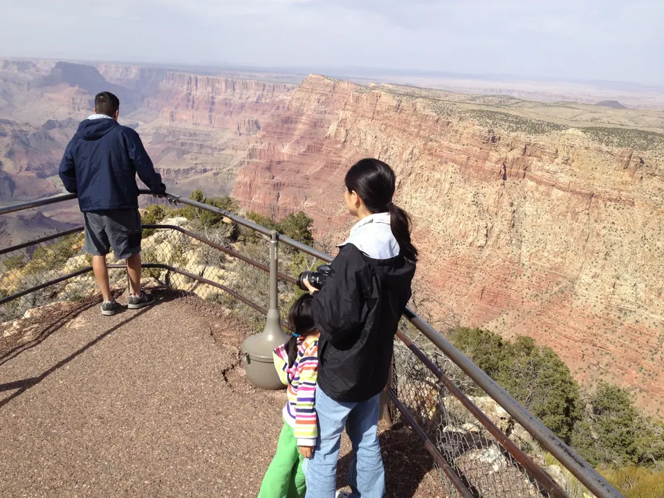 A man in a navy jacket looks out over the railing while a woman in a black jacket holding a camera and a girl in a striped jacket and green pants stand beside him, with the red canyon wall opening behind.
