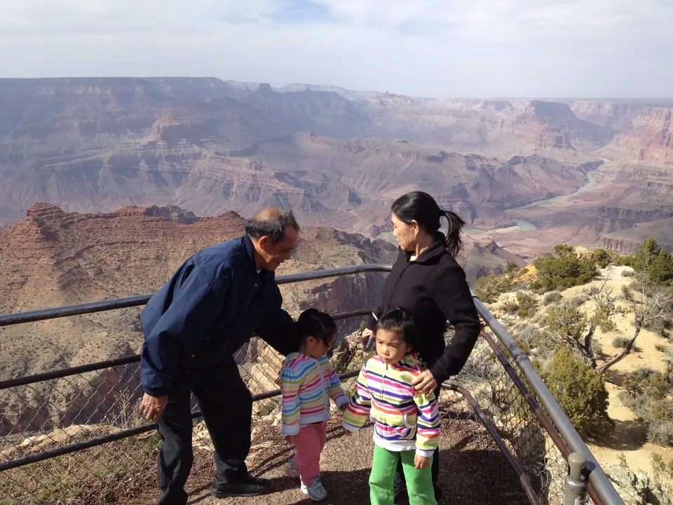 An older man in a navy jacket leans toward a small girl in a striped jacket while a woman in black stands with another girl in green pants, at a railing above the canyon and river.