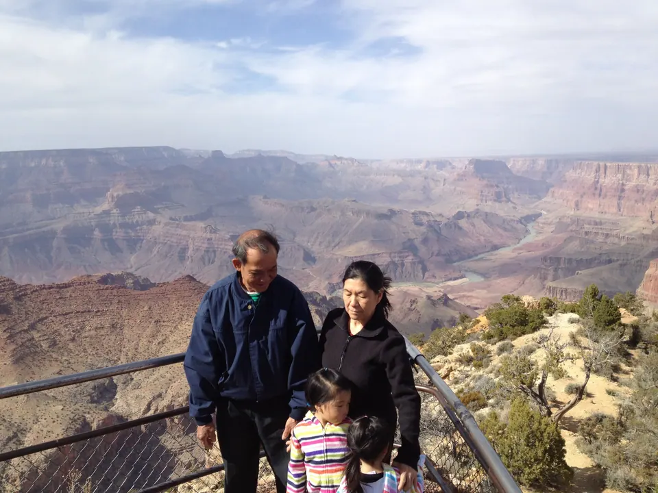 An older man and an older woman in dark jackets pose at the rail with two girls in striped jackets in front of them, the canyon and the river below and a hazy sky.