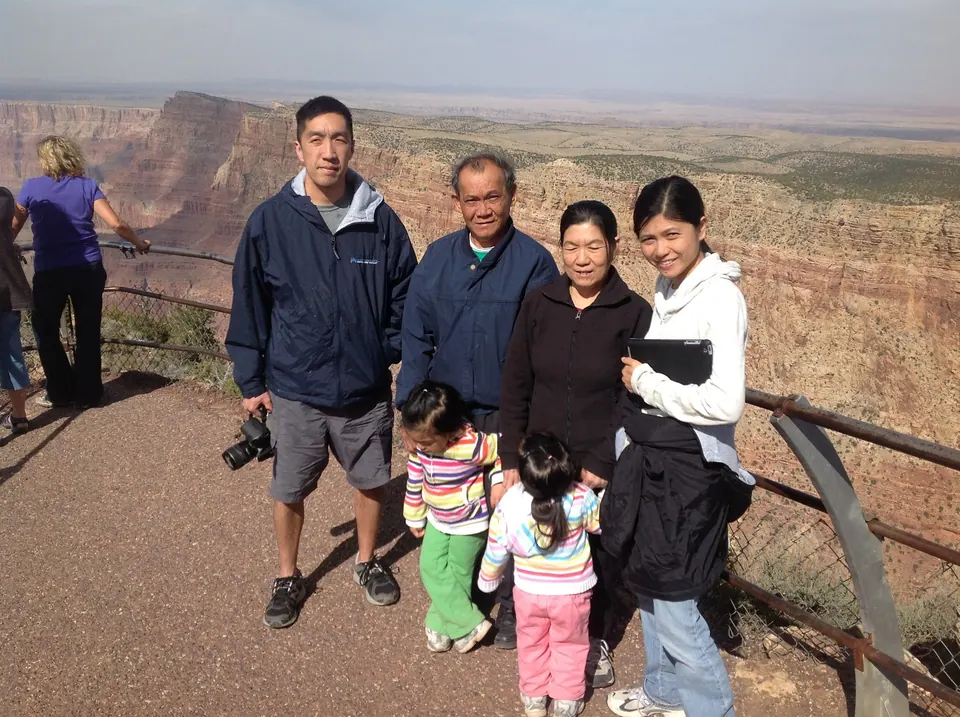 Four adults and two small girls stand at a railing on the canyon rim, with layered cliffs and a flat plateau stretching behind them.