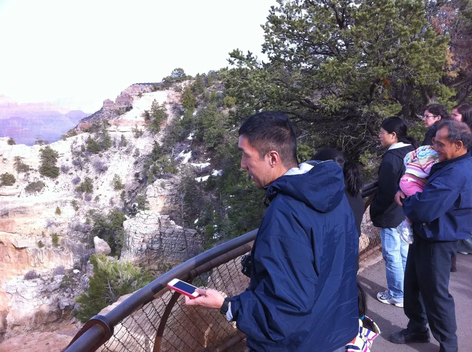 A man in a navy jacket holds up a pink phone at the railing while other visitors, one carrying a baby, look out at a snow-dusted rim with pines.
