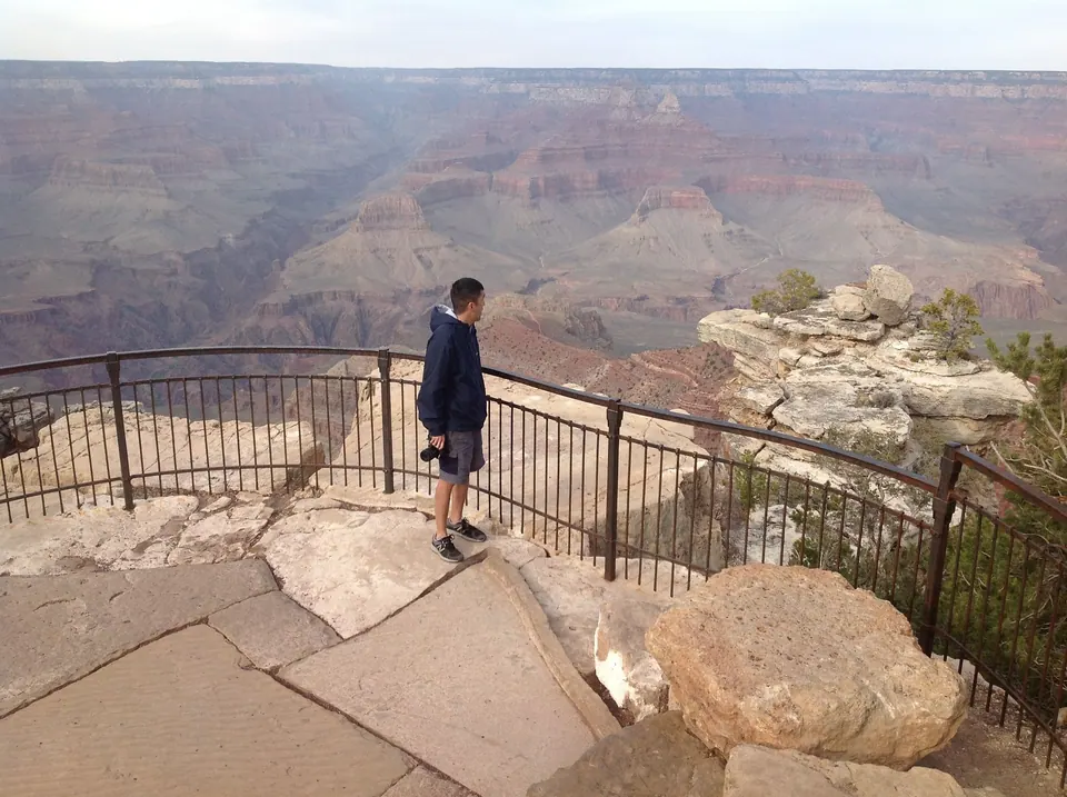 A man in a navy jacket with a camera in his hand stands at an iron railing looking out at a hazy canyon and a rock outcrop.