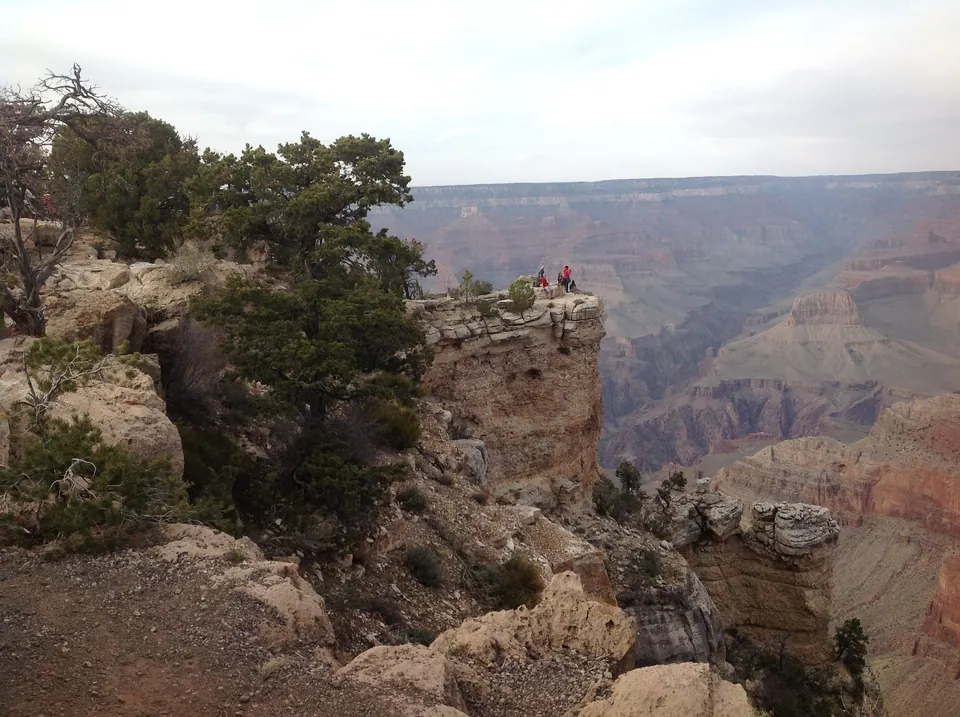 A pinnacle of banded rock with twisted pines juts into the canyon, with a few tiny visitors standing on a far viewpoint.