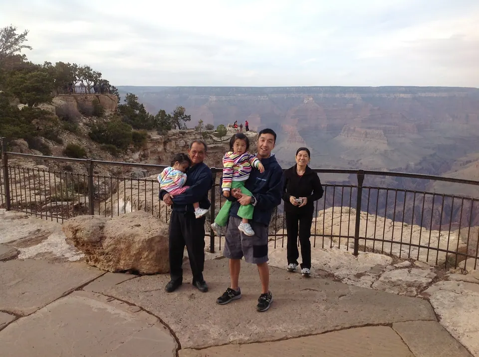 Against an iron railing, an older man in dark clothes carries a sleepy girl, a man in a navy jacket carries a girl in a striped jacket and green pants, and a woman in black stands at the right, the canyon fading in the dusk behind.
