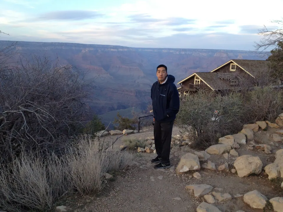 A young man in a dark hooded jacket stands on a rocky path at the canyon rim at dusk, with a gabled wooden building to one side and the canyon fading blue behind.