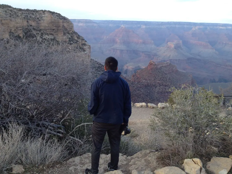 Seen from behind, a man in a navy jacket with a camera looks out over the darkening canyon from a rocky path with dry brush.