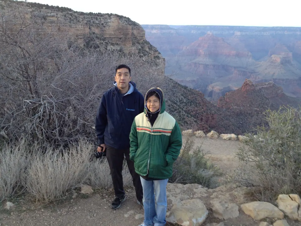A man in a navy jacket with a camera stands beside a woman in a green hooded jacket on the rim path, with the canyon and a red butte behind them.
