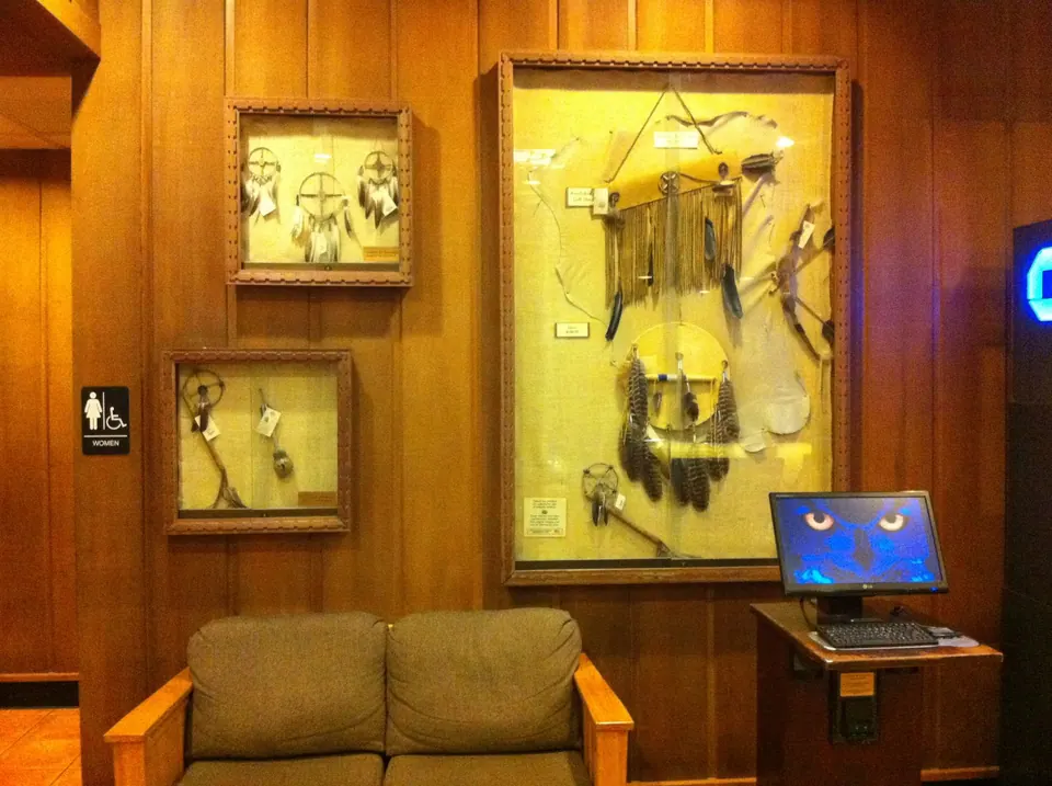 A wall of Native American dreamcatchers and feather displays in glass cases above a two-seat sofa and a computer kiosk, in a wood-paneled lobby.