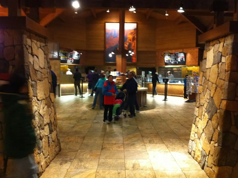 A tiled hall with stone pillars and a canyon painting over serving counters, where a few visitors and a stroller wait in line.