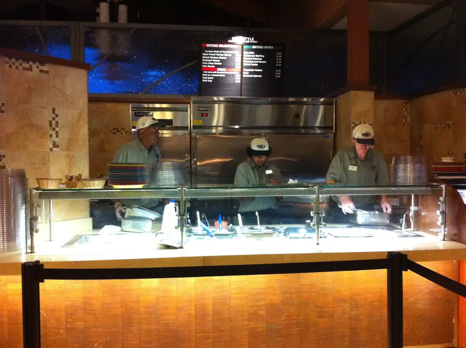 Three staff in green shirts and caps work behind a glass-fronted serving counter under a menu board of entrees and sides.