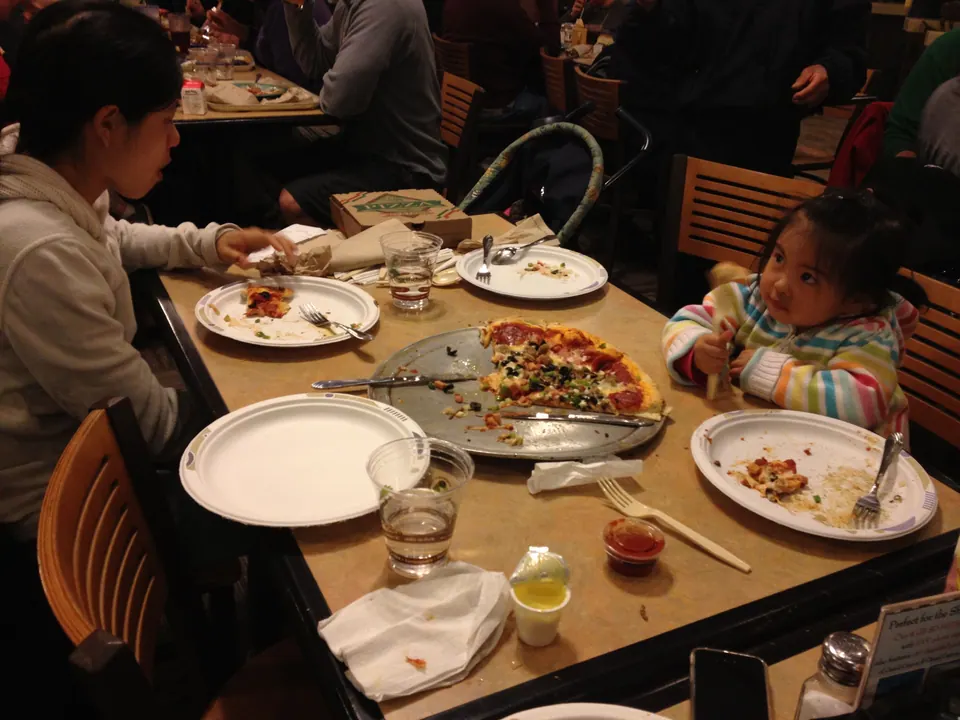 A little girl in a striped jacket looks up from her paper plate at a table with a pizza tray, while a woman eats across from her.