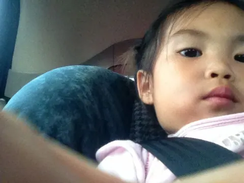 A close, grainy shot of a little girl in a pink top buckled into a car seat, looking at the camera.