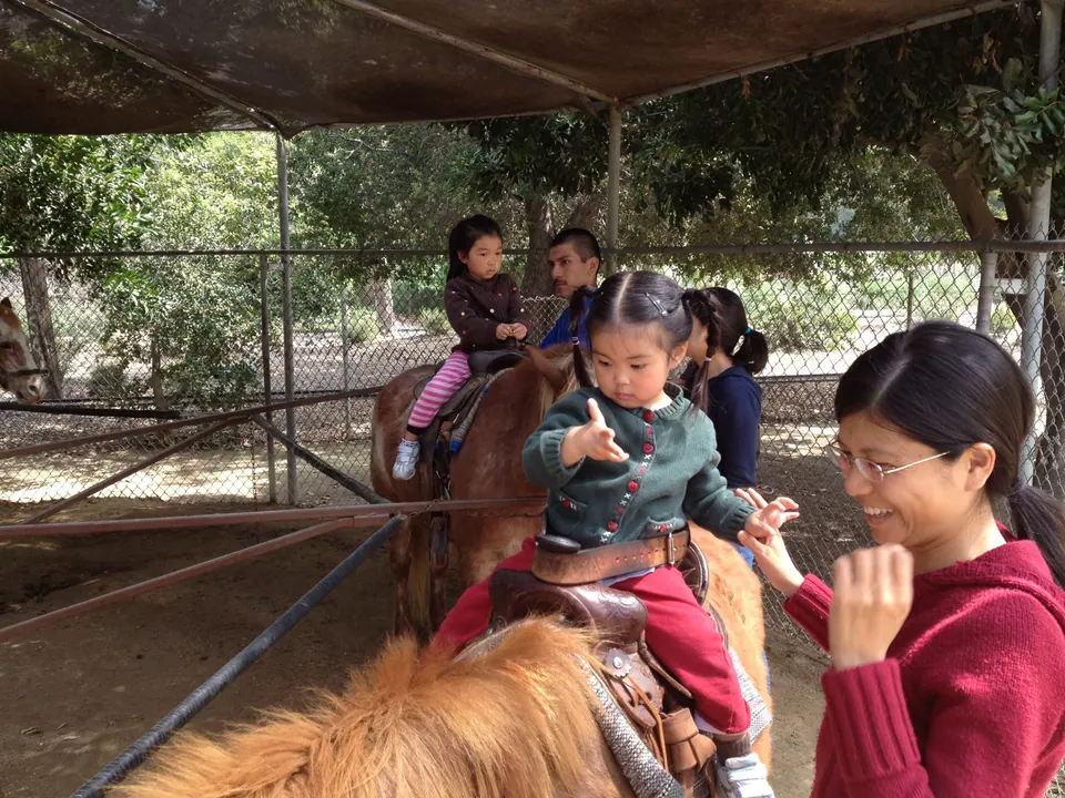 Under a shade roof, a little girl in a green jacket rides a pony and reaches toward a smiling woman in red; another small girl rides a second pony behind, led by a man.