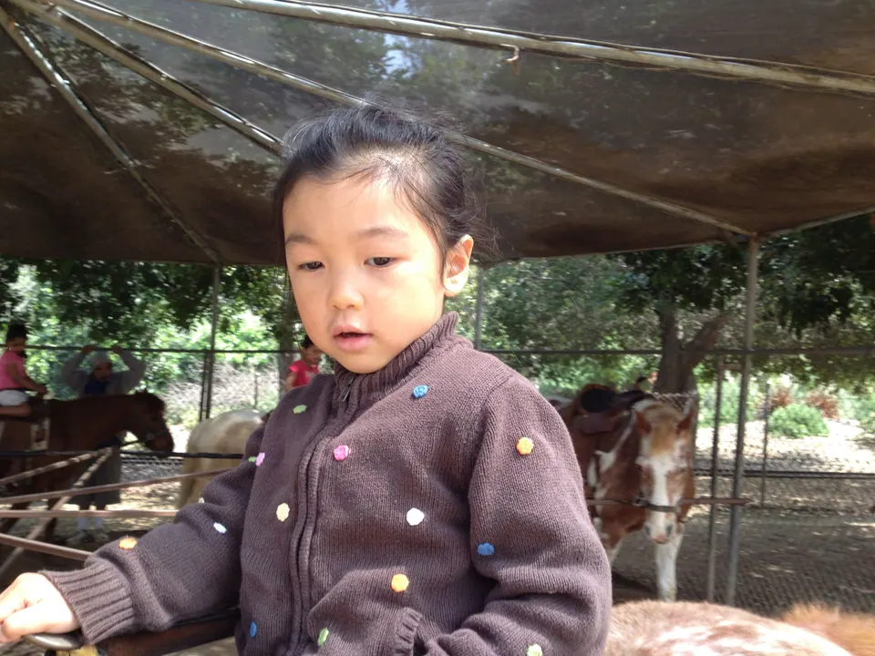 A close view of a girl in a brown cardigan with colored dots, mouth open as if speaking, on her pony under the shade roof with other ponies behind.