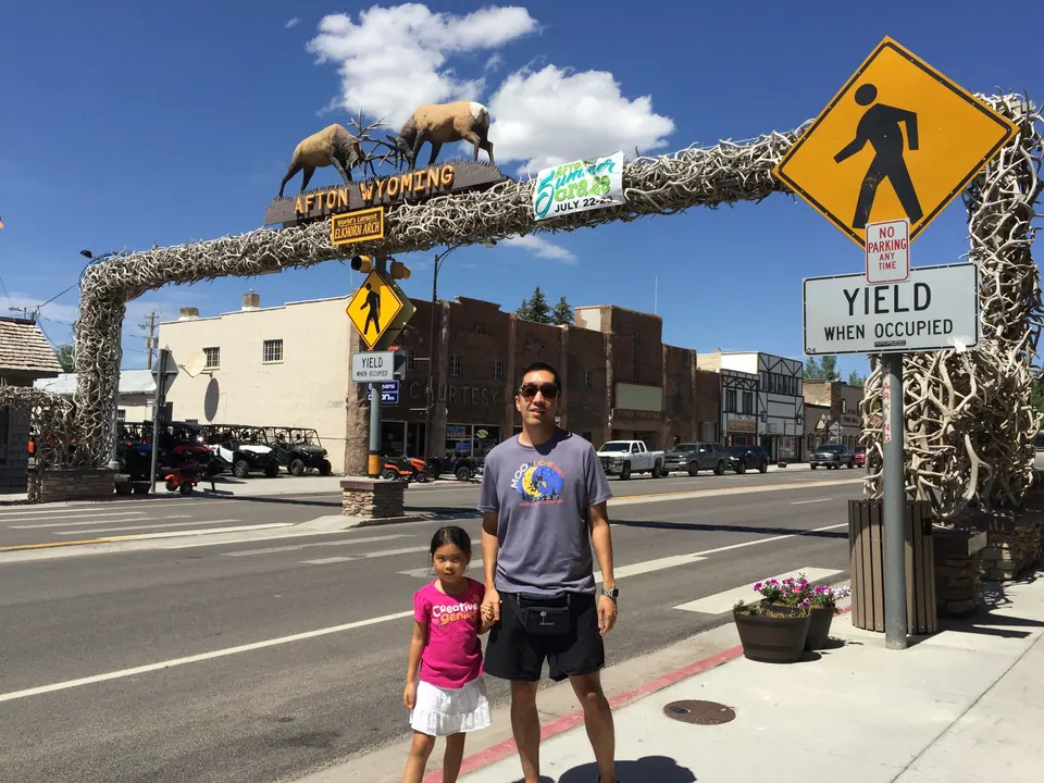 A man in sunglasses and a small girl in pink stand on a sidewalk beneath a tall arch of piled elk antlers; a sign on top reads Afton Wyoming, with two elk statues above it.