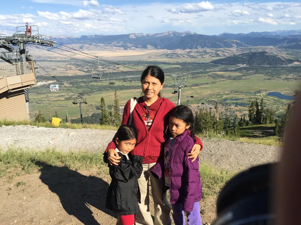 A woman in a red jacket hugs two girls on a gravel summit beside a lift station, with chairlifts, a broad green valley and distant mountains behind them.