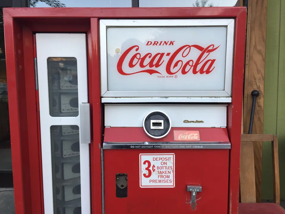 A red vintage Coca-Cola bottle vending machine with a sign about a three-cent deposit on bottles.