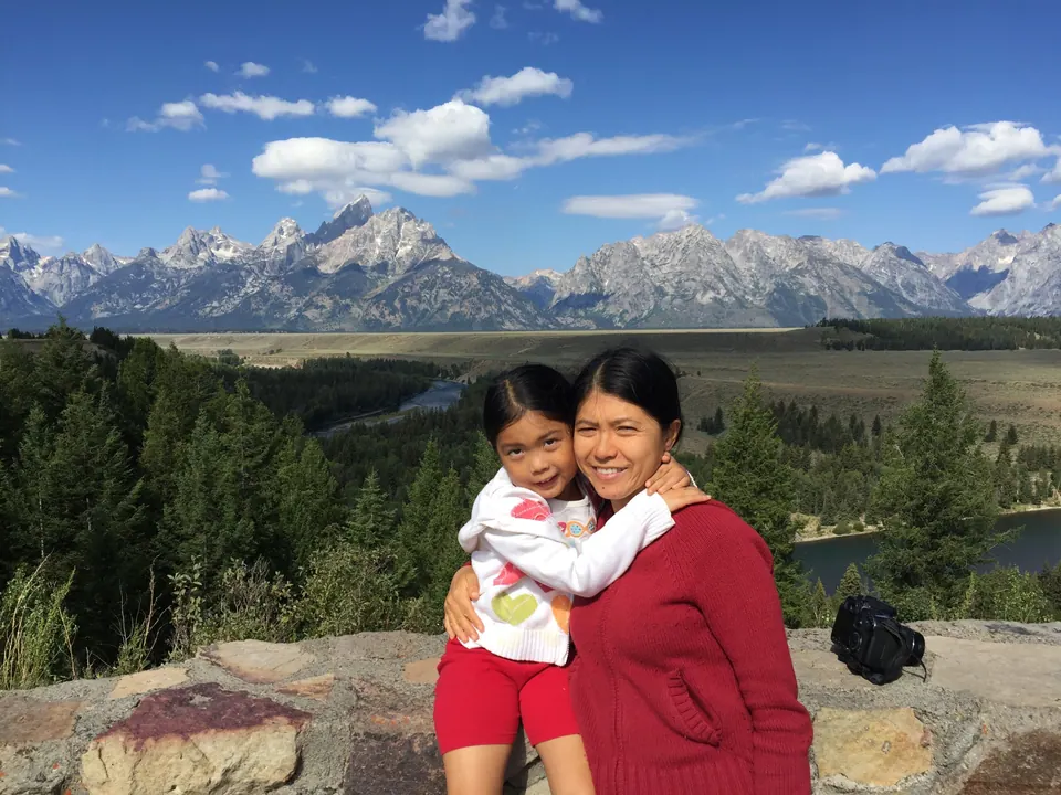 A woman in a red sweater hugs a small girl on a stone wall; behind them a forested river valley runs up to a long line of jagged gray peaks under a blue sky.