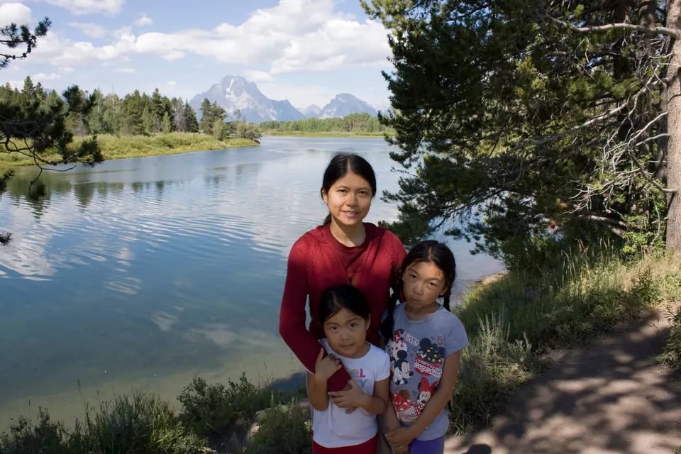 A woman in red stands with two girls on a riverbank path; a calm river and a snowy mountain are behind them.