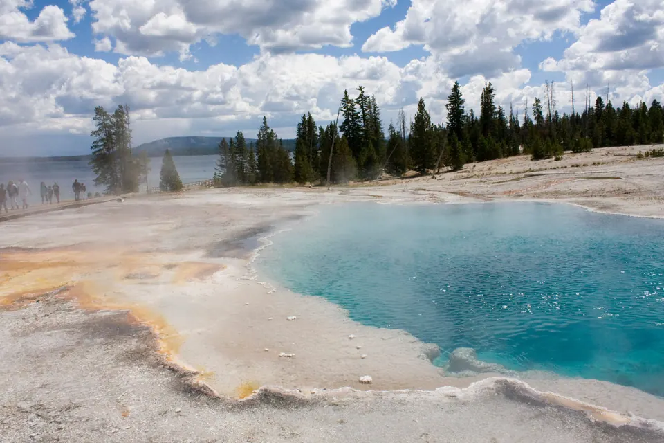 A deep turquoise hot pool with orange edges on a flat white crust, pine trees and a large lake behind under towering clouds; people walk along a boardwalk at the left.