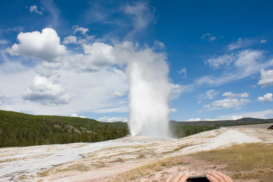 A tall white column of water and steam erupts from a pale geyser mound against a deep blue sky with clouds; a spectator's hands hold up a phone at the bottom edge.