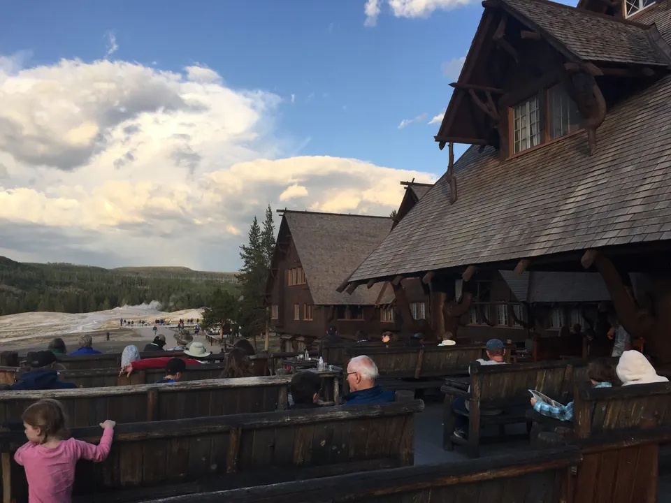 People sit on wooden benches on a large deck beside a steep shingled log building; a child in pink leans on the rail, and steam rises from a pale geyser basin under evening clouds.