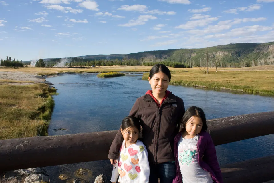 A woman and two girls stand at a log railing on a bridge over a clear river; steam rises from the far bank under a pale blue morning sky.