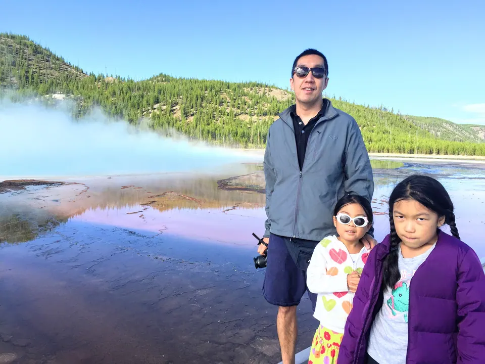 A man in sunglasses and a gray jacket stands with two girls beside a vast, shallow hot spring streaked with orange and blue, steam drifting across it and a forested hill behind.