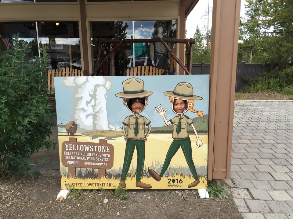 Two children's faces peek through a painted board of two cartoon park rangers beside Old Faithful and a sign reading Yellowstone, celebrating 100 years with the National Park Service.