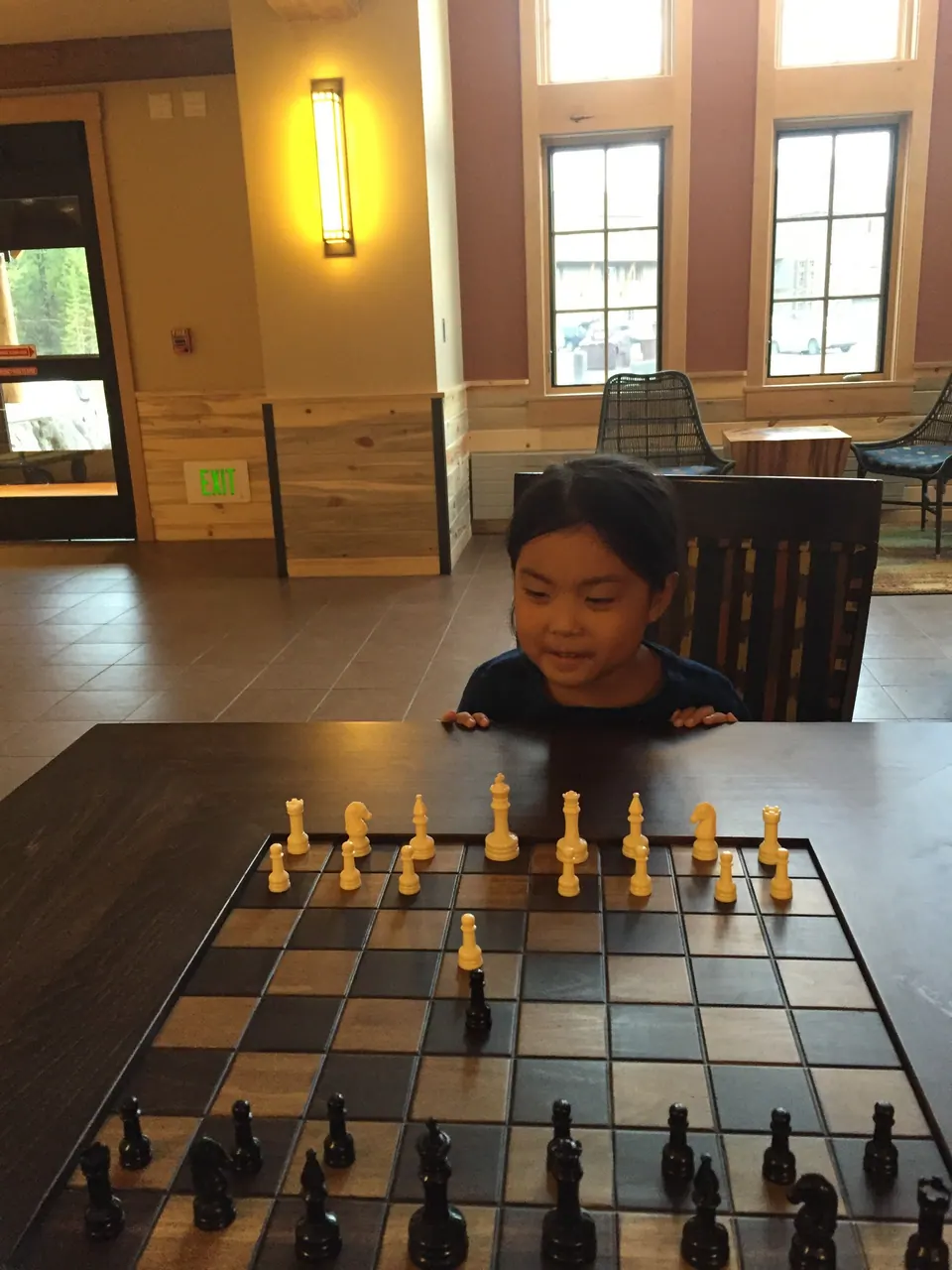 A smiling girl peeks over the edge of a large wooden chessboard set up on a table in a bright lobby with tall windows.