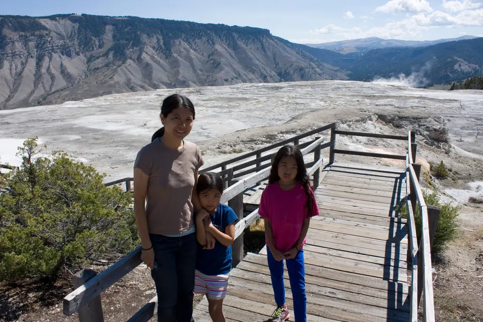 A woman and two girls stand on a wooden boardwalk staircase above pale, steaming terraces, with a steep gray ridge behind them.