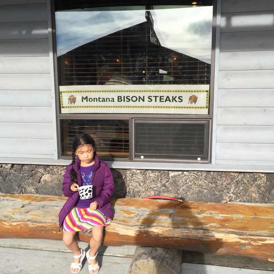 A girl in a purple jacket sits on a log bench holding a cup, below a restaurant window with a sign reading Montana Bison Steaks.