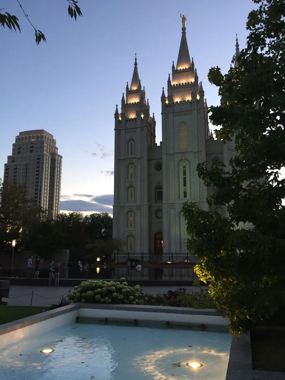 The floodlit granite spires of a large temple against a violet dusk sky, with a lit fountain pool in the foreground and a high-rise at the left.