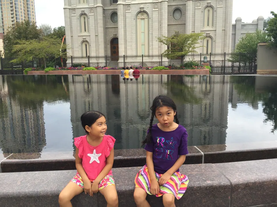 Two girls sit on the stone rim of a long reflecting pool; a gray granite temple and its reflection fill the background on an overcast morning.