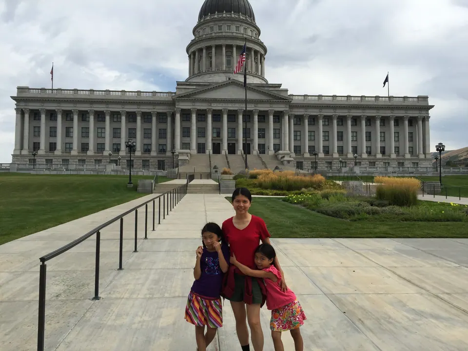 A woman in red hugs two girls on a wide walkway below a domed, columned capitol building under a gray sky.