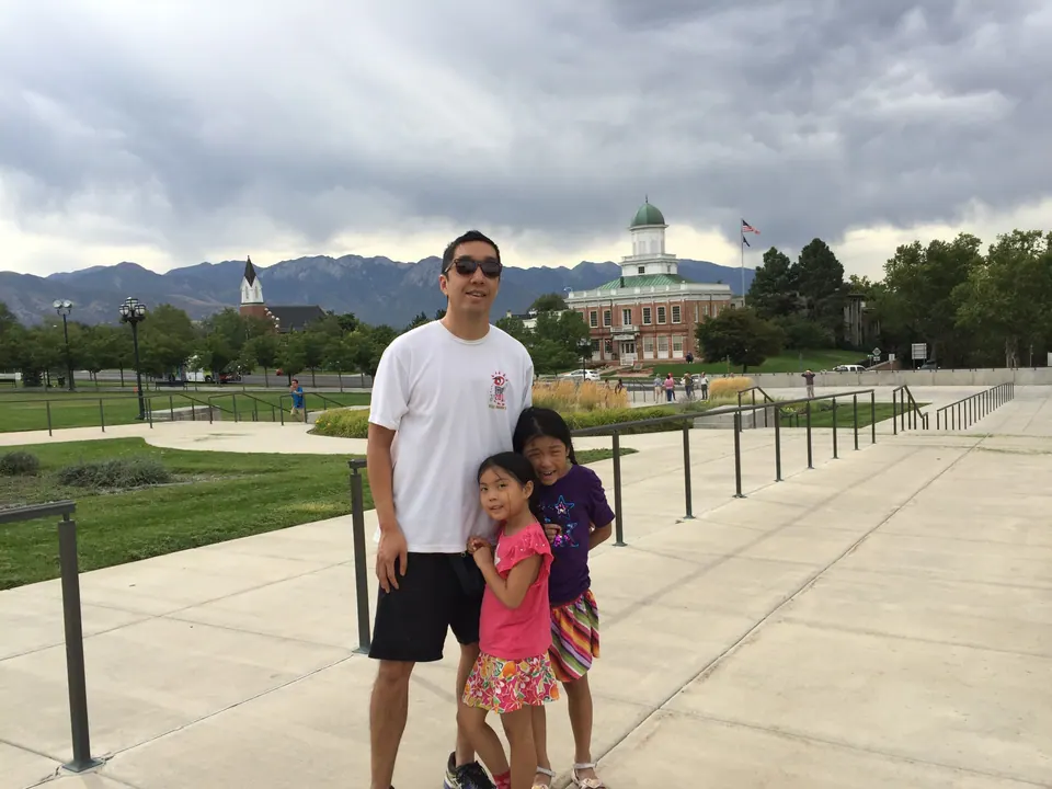 A man in sunglasses and a white T-shirt stands with two girls on a paved plaza; behind them are a red-brick building with a green dome, a steeple and dark clouds over a mountain range.